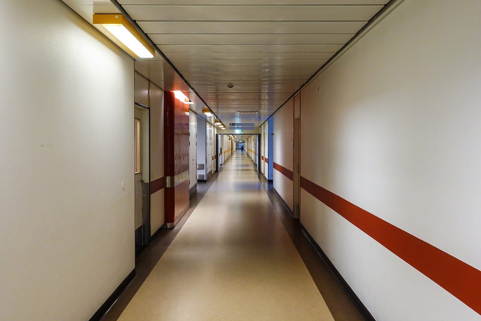 A long, empty hospital corridor with closed doors and a red band painted along one wall