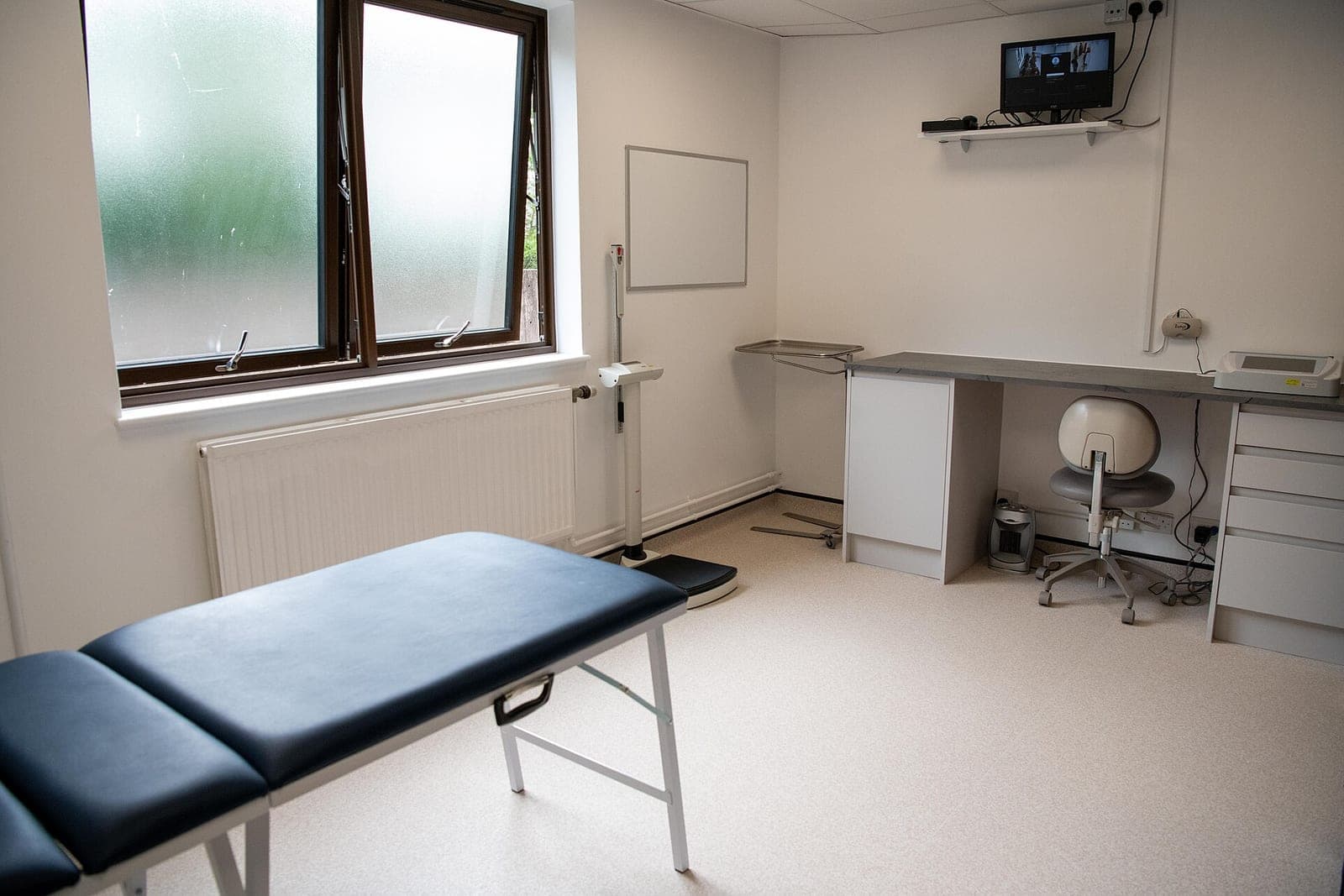 An empty exam room with a padded treatment table, a frosted window, a height scale and a corner desk