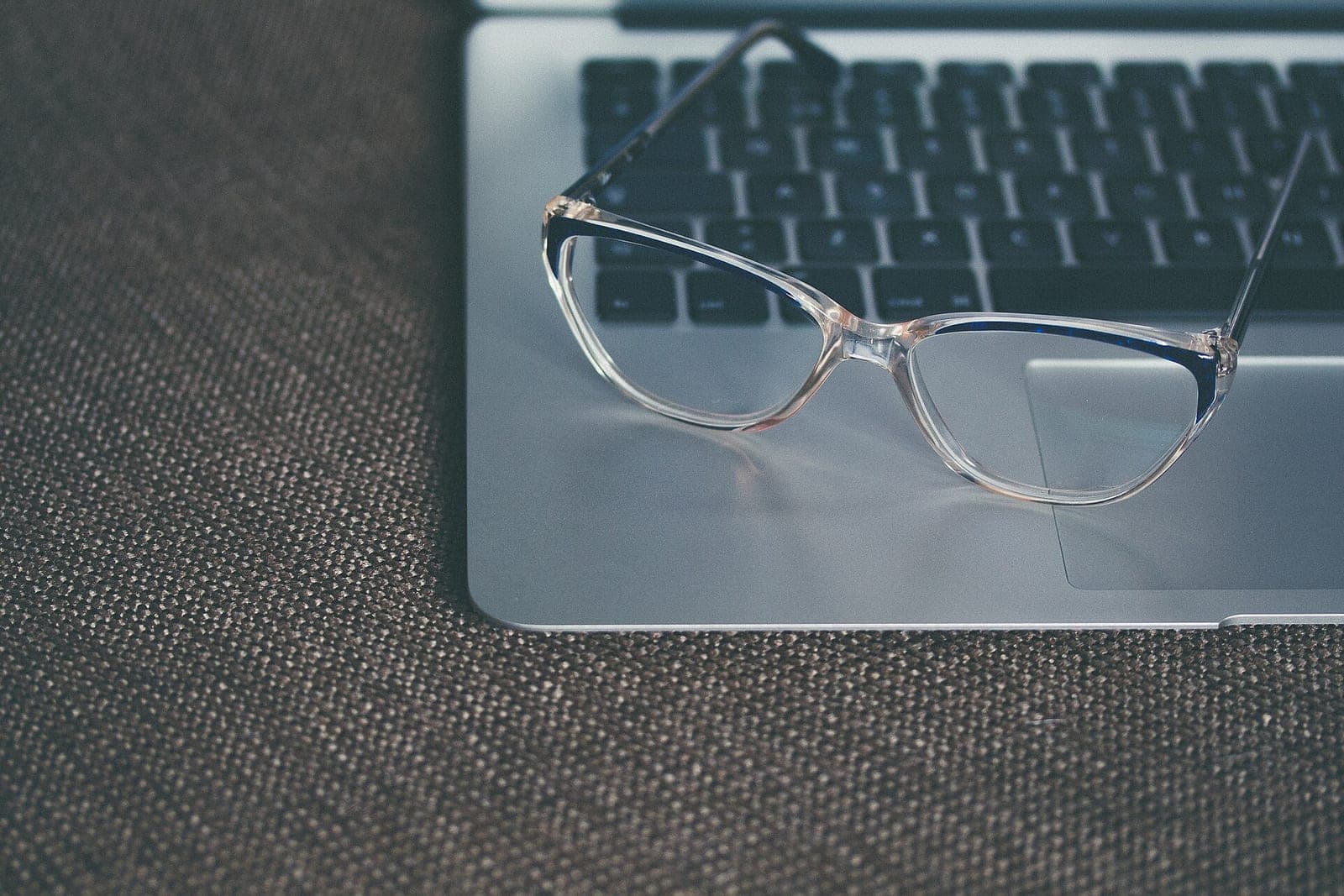 A pair of clear-framed glasses resting on an open silver laptop on a brown woven cloth