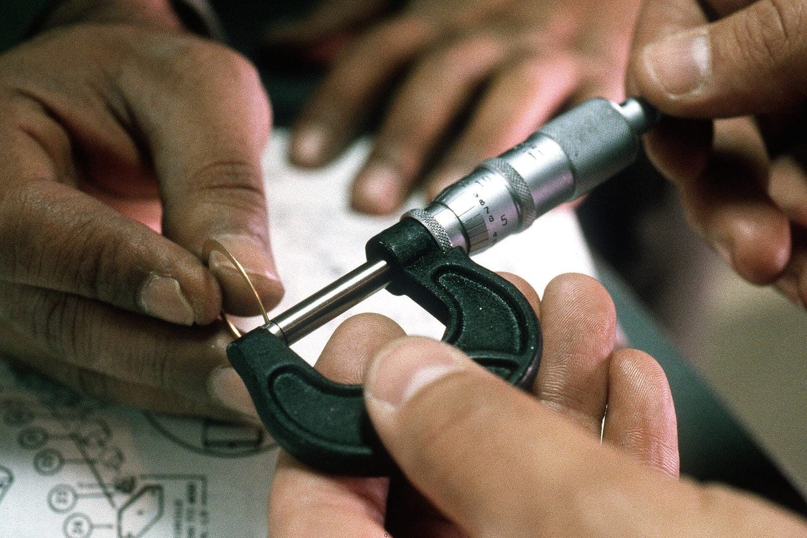 Two pairs of hands hold a micrometer against a thin metal ring above a printed drawing