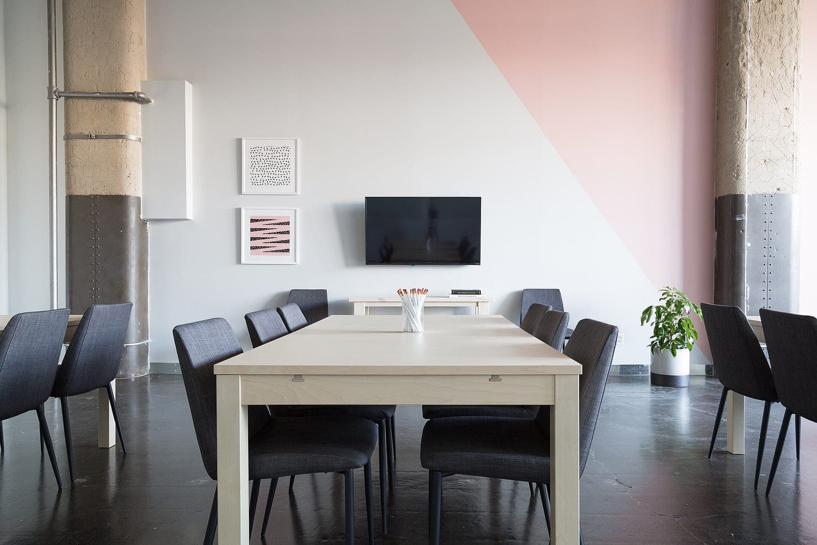 An empty meeting room with a long pale wood table, grey chairs, a wall screen and a potted plant by a pink wall