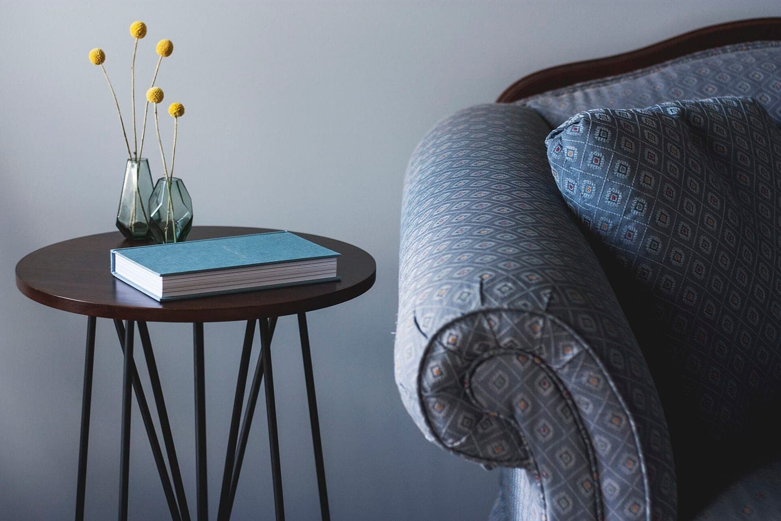 A side table with a blue book and a vase of yellow flowers beside a patterned armchair