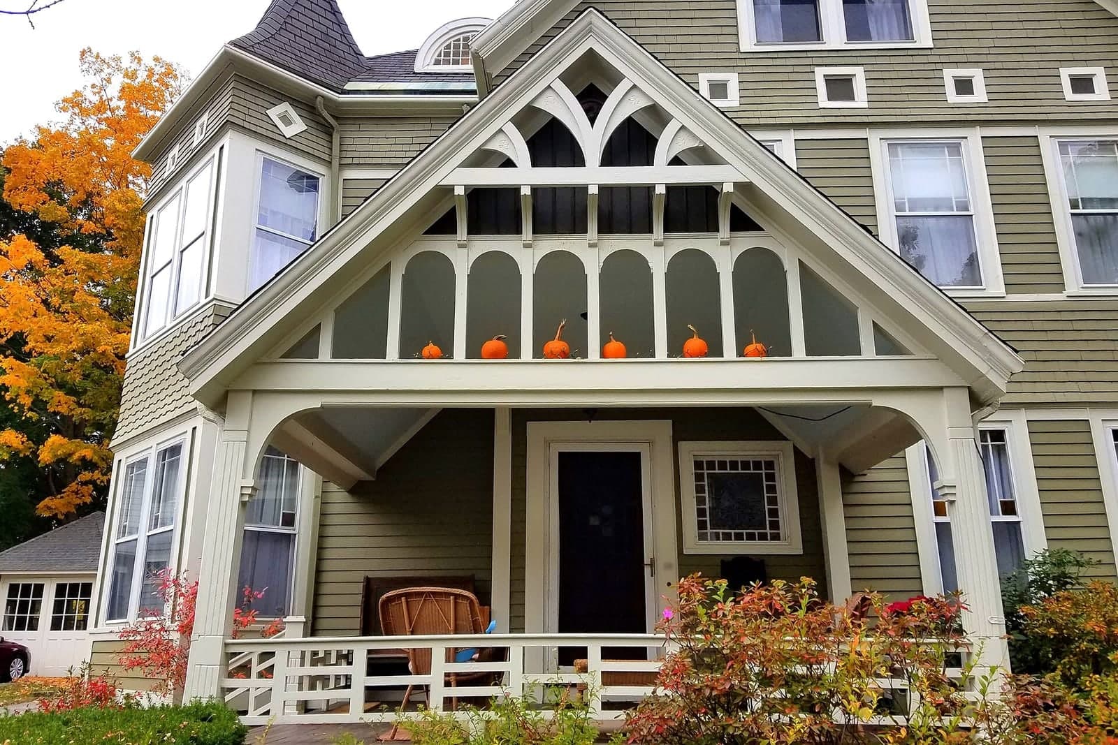 A green clapboard house with a covered front porch, small pumpkins along the gable and an orange maple beside it
