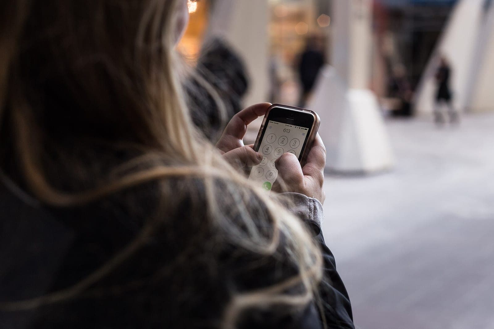 Seen over a person's shoulder, a hand taps a phone keypad in a busy indoor walkway