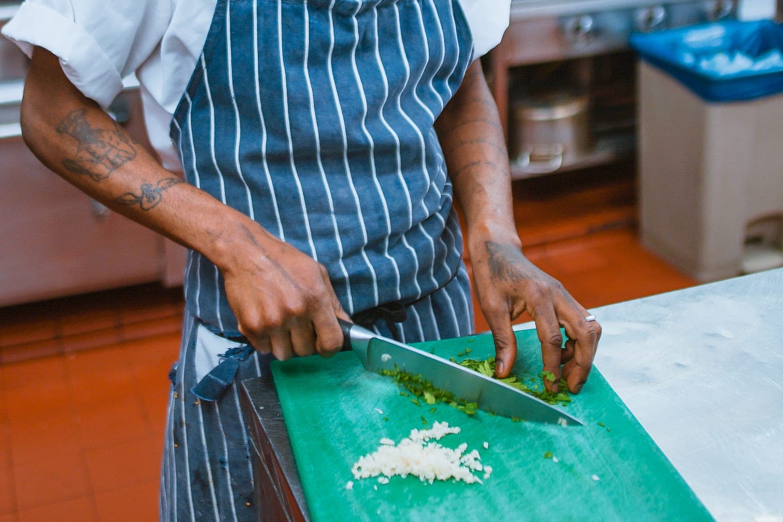 A cook in a striped apron chops green herbs on a green cutting board at a steel prep counter, with a range and stockpots behind