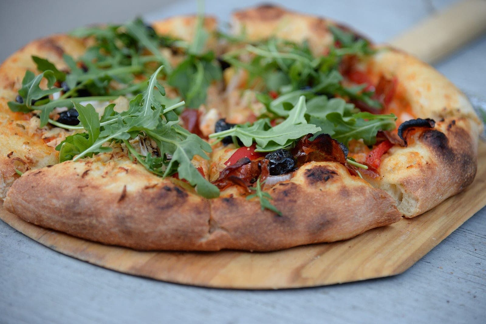 A freshly baked pizza topped with arugula, olives and peppers on a wooden pizza peel