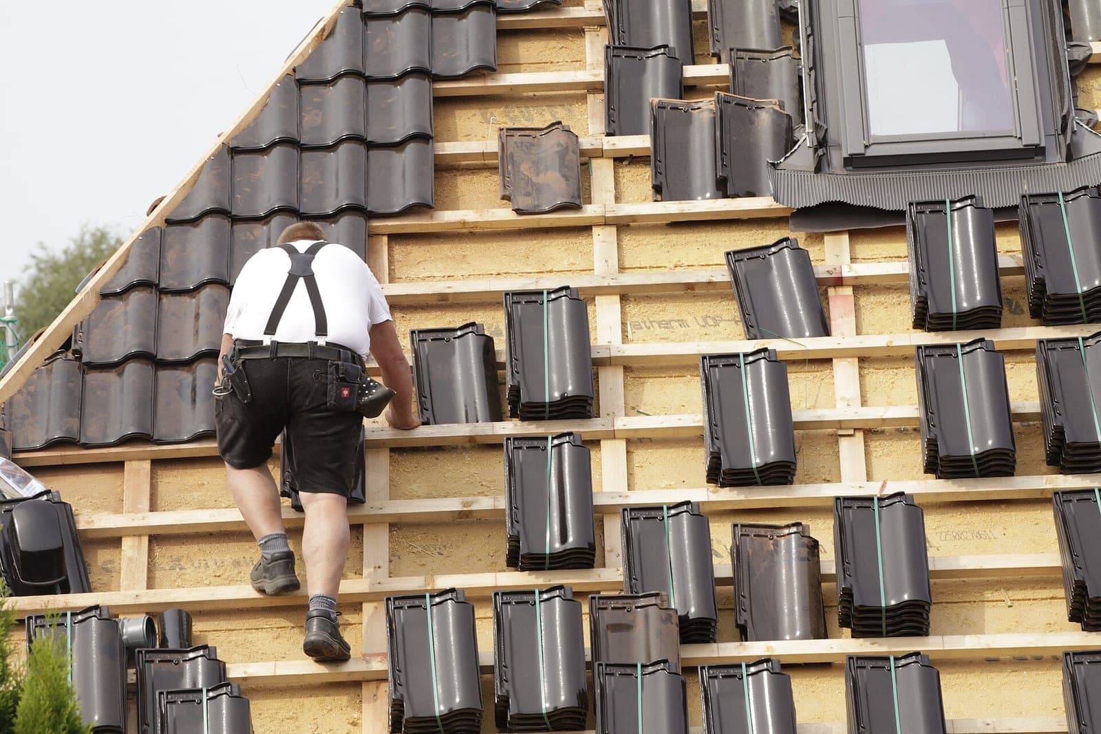 A roofer seen from behind climbs roof battens past stacks of dark clay tiles