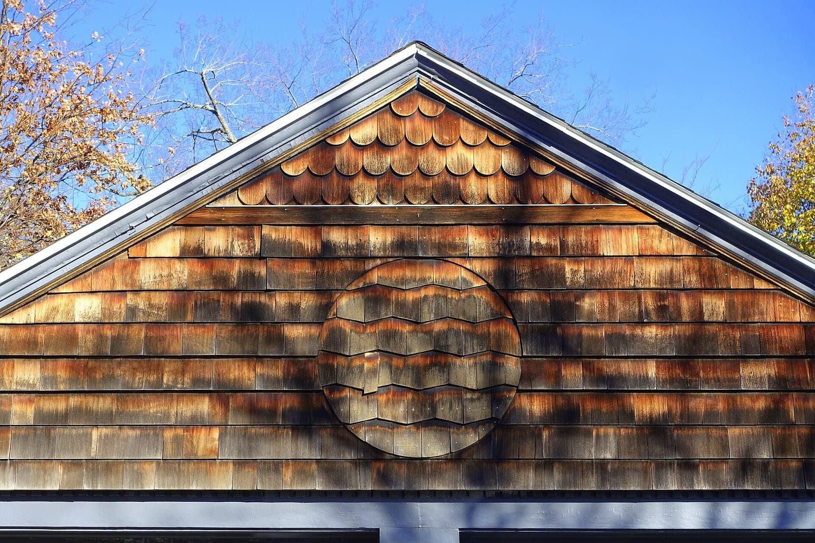 A wood shake gable with a round shingle pattern under a blue autumn sky