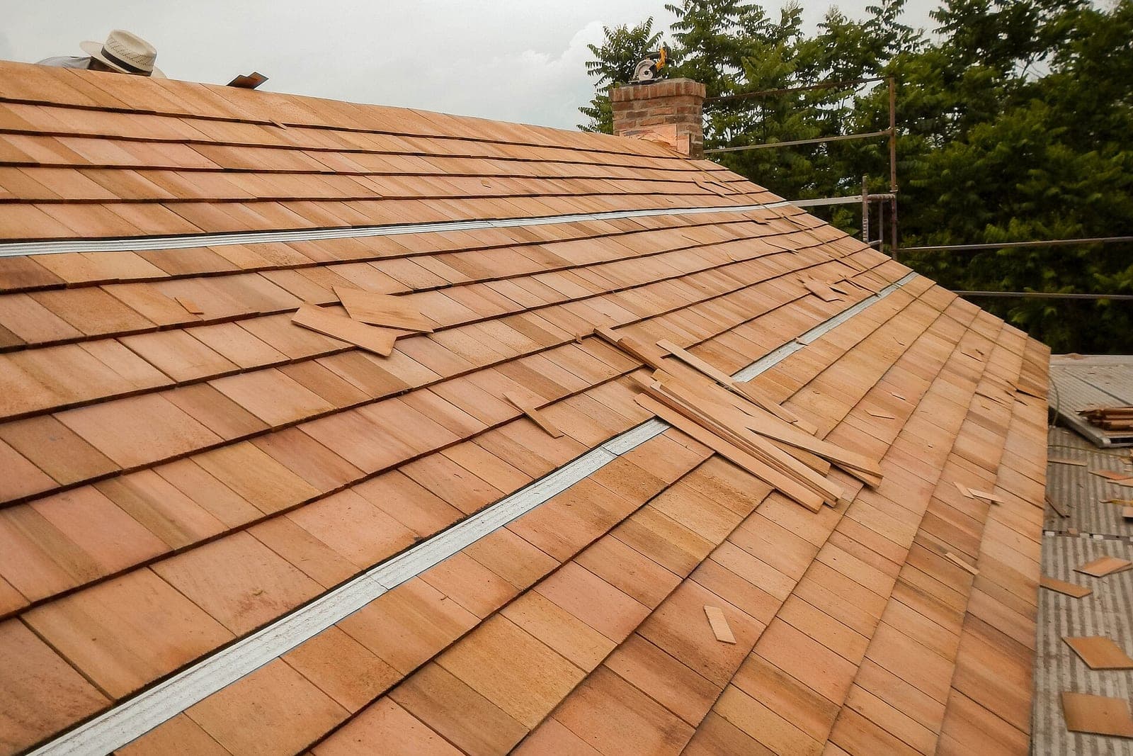 A nearly finished cedar shingle roof slope with loose shingles stacked near a brick chimney