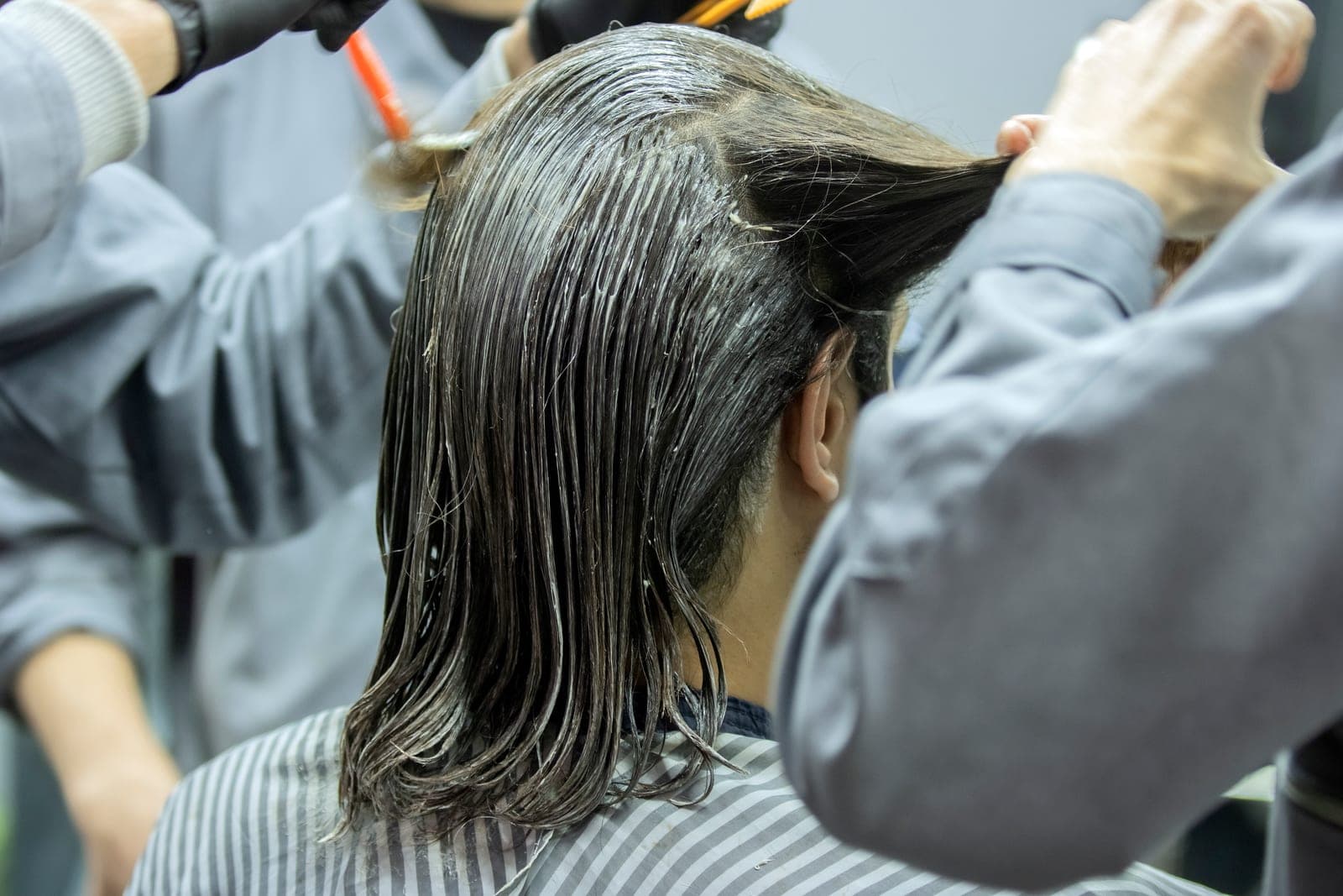 Seen from behind, a client's wet, color-coated hair is lifted by one stylist's bare fingers while another's gloved hands hold a tint brush