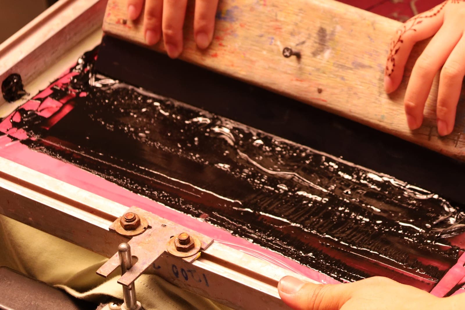 Two hands pull a worn wooden squeegee through thick black ink across a pink screen in a metal frame