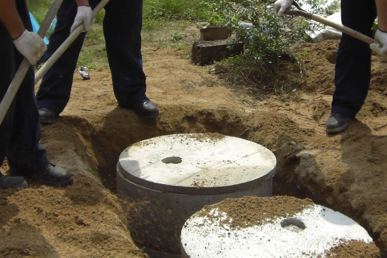Workers with shovels stand around two round concrete septic tank lids set in fresh dirt in a green yard