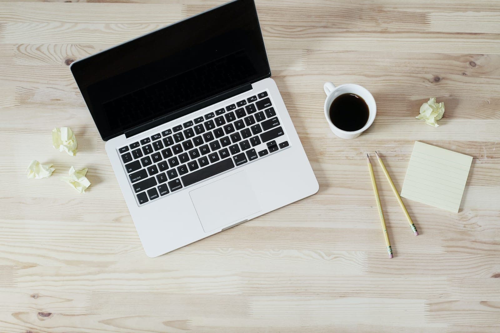 A laptop on a light wooden desk next to a cup of coffee, two pencils and a sticky note