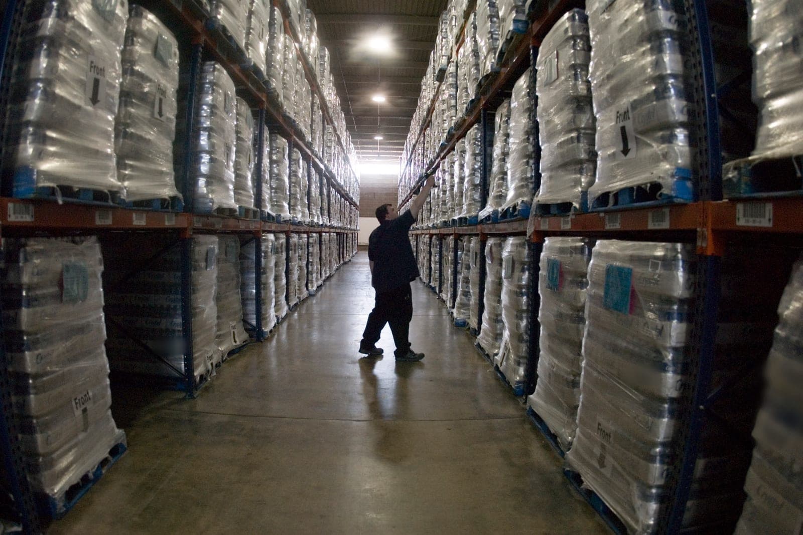 A worker reaches up to a pallet in a tall warehouse aisle lined with wrapped stock