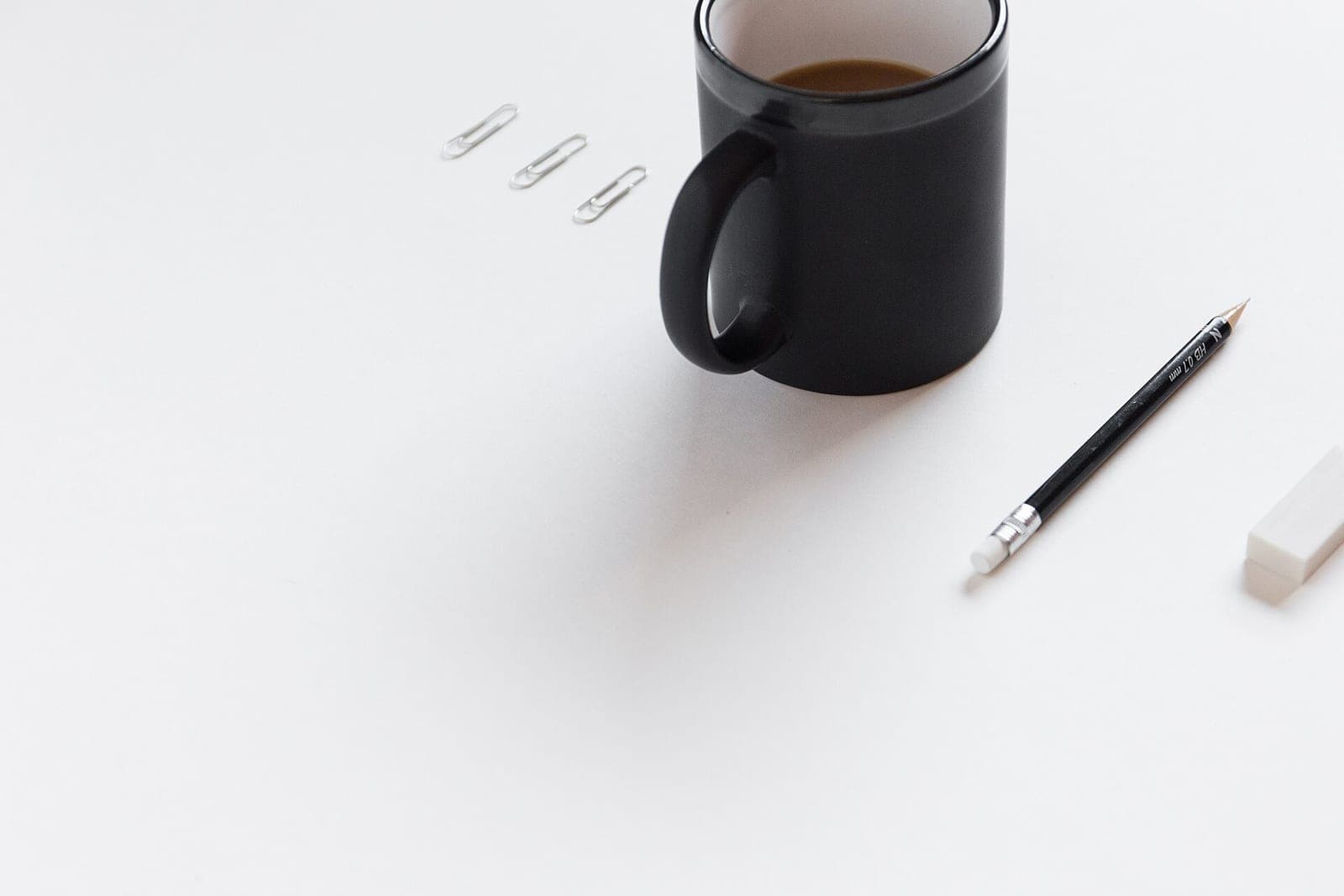 A black coffee mug, a pencil, an eraser and three paper clips on a clean white desk