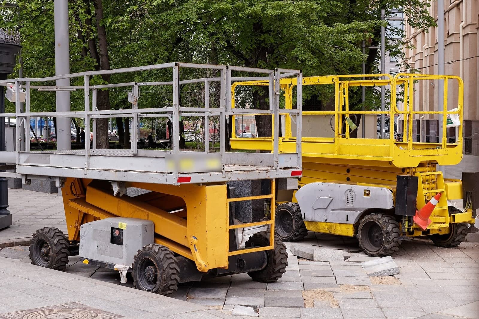 Two scissor lifts, one gray and one yellow, parked side by side on a paved city sidewalk