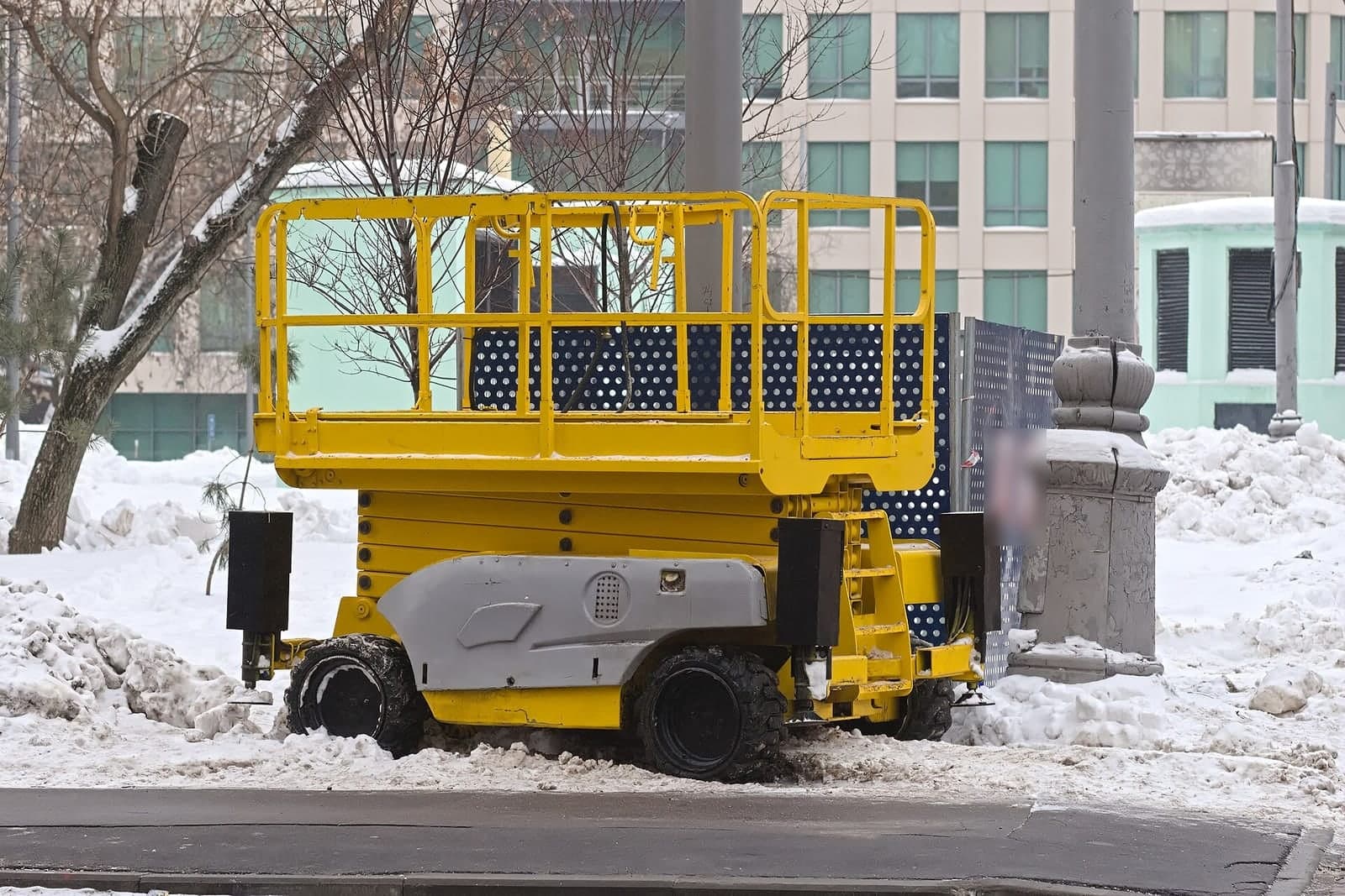 A yellow scissor lift parked in the snow beside a city street