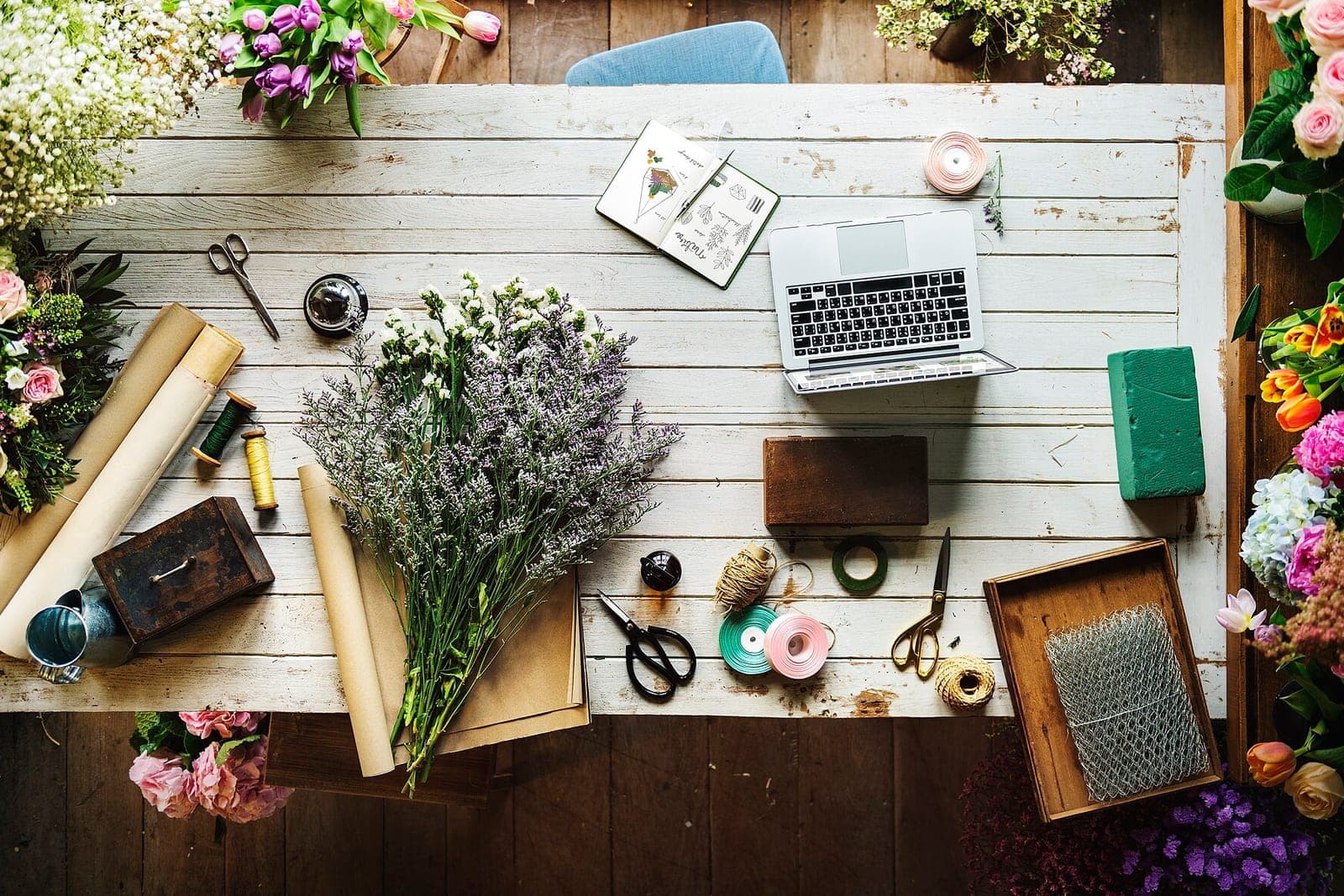 A florist's white worktable with bunches of lavender, ribbon spools, scissors, a sketchbook and a laptop