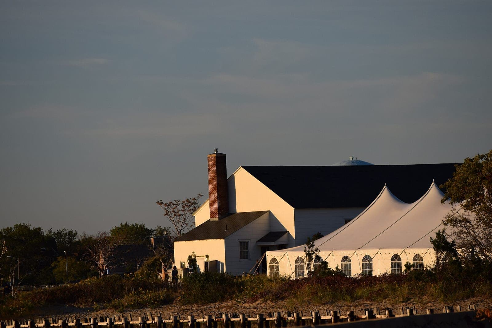 A white peaked event tent stands beside a pale wooden building in low evening sun