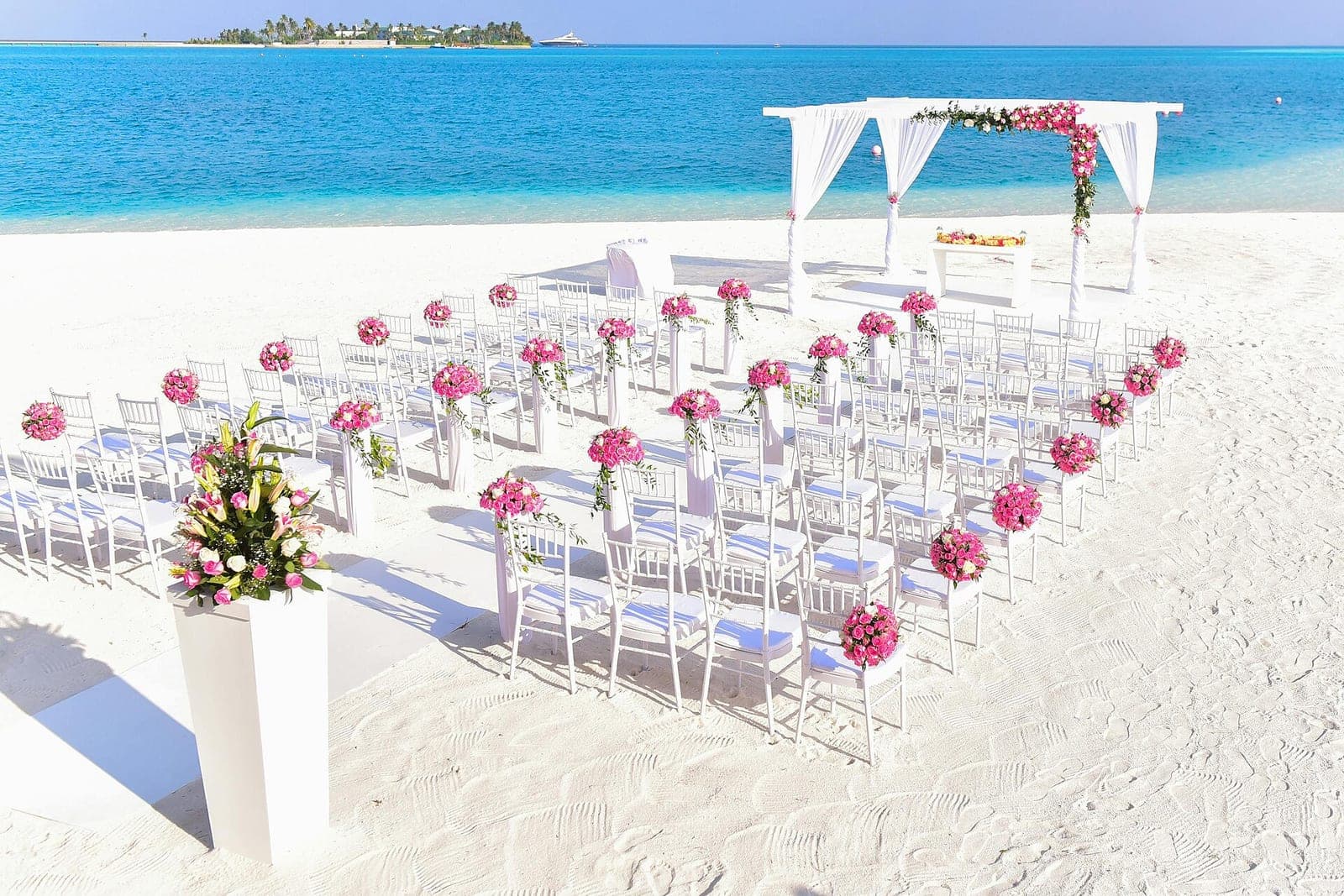 White chairs, pink flower posts and a draped arch set up on an empty beach before a ceremony