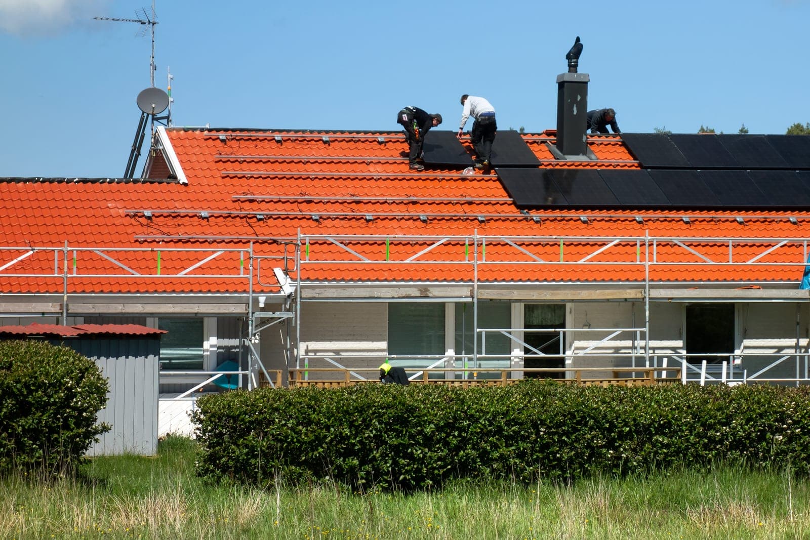 A crew fits black solar panels onto an orange tile roof above scaffolding on a sunny day