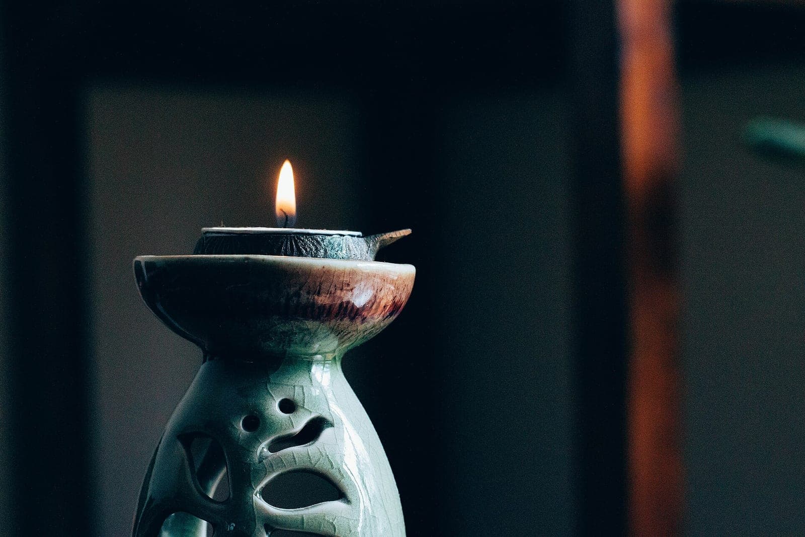 A small candle burns on top of a pale green ceramic oil warmer in a dim room