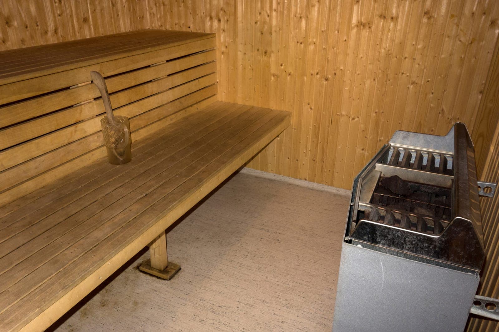 A wooden sauna room with slatted benches, a ladle in a glass cup and a steel heater full of stones