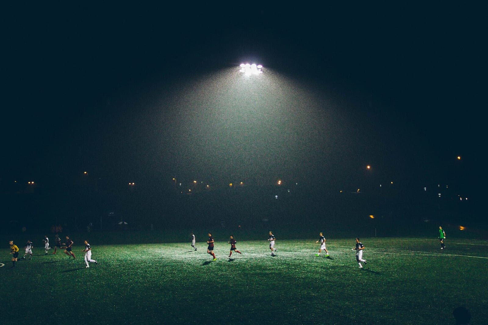 Adult players in matching kits run across a turf field at night under one bright light tower