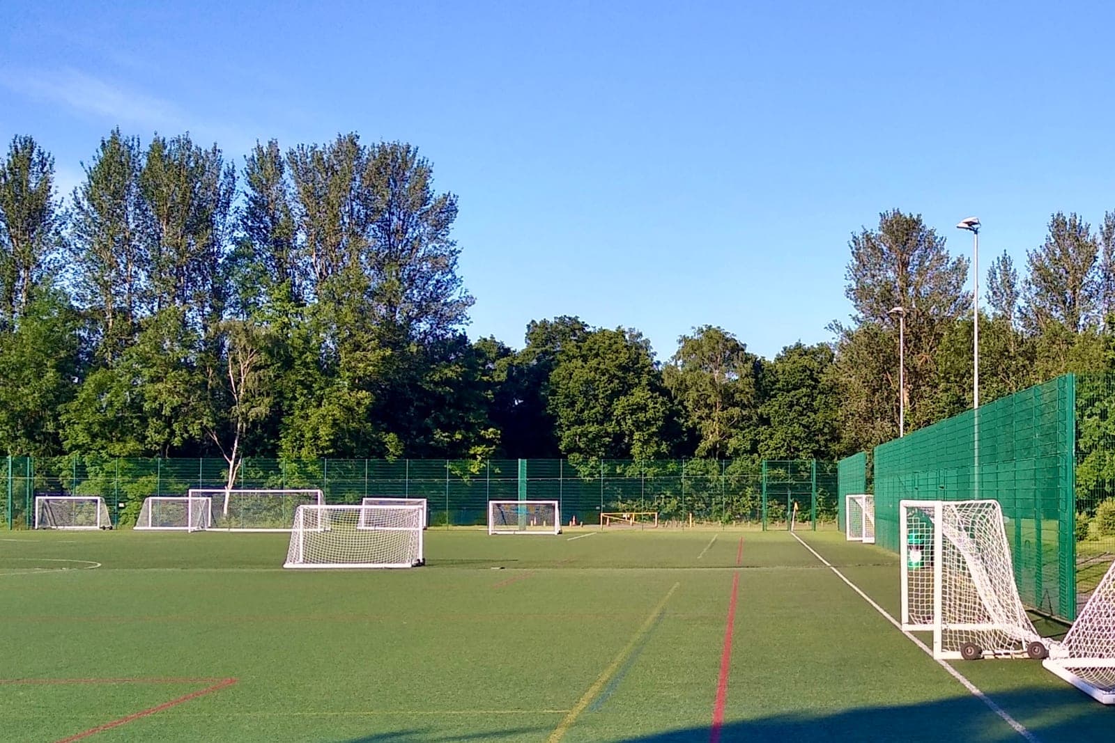 White goals of several sizes stand around an empty artificial turf field inside a tall green fence