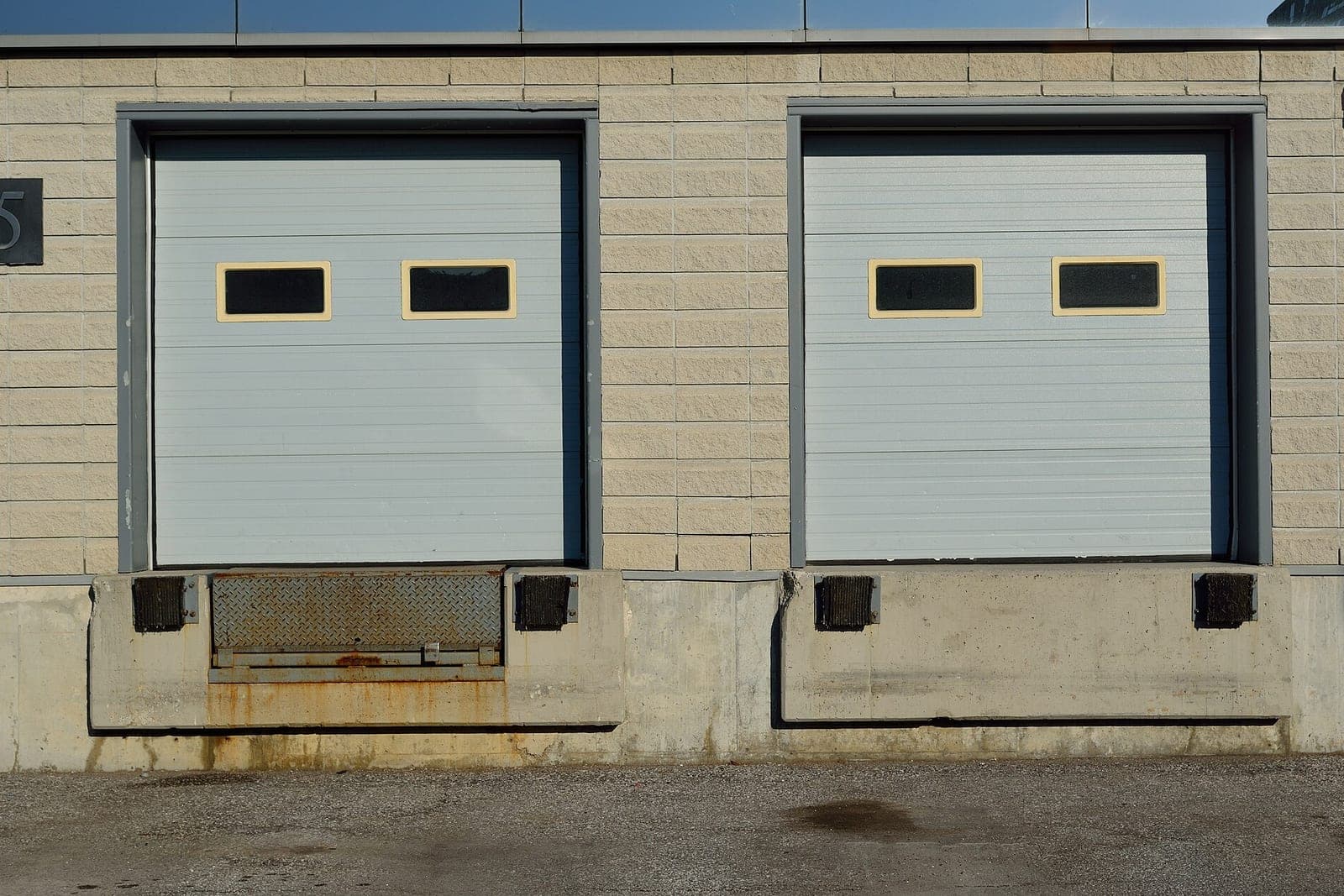 Two closed white loading dock doors with small windows in a beige block wall above concrete dock bays