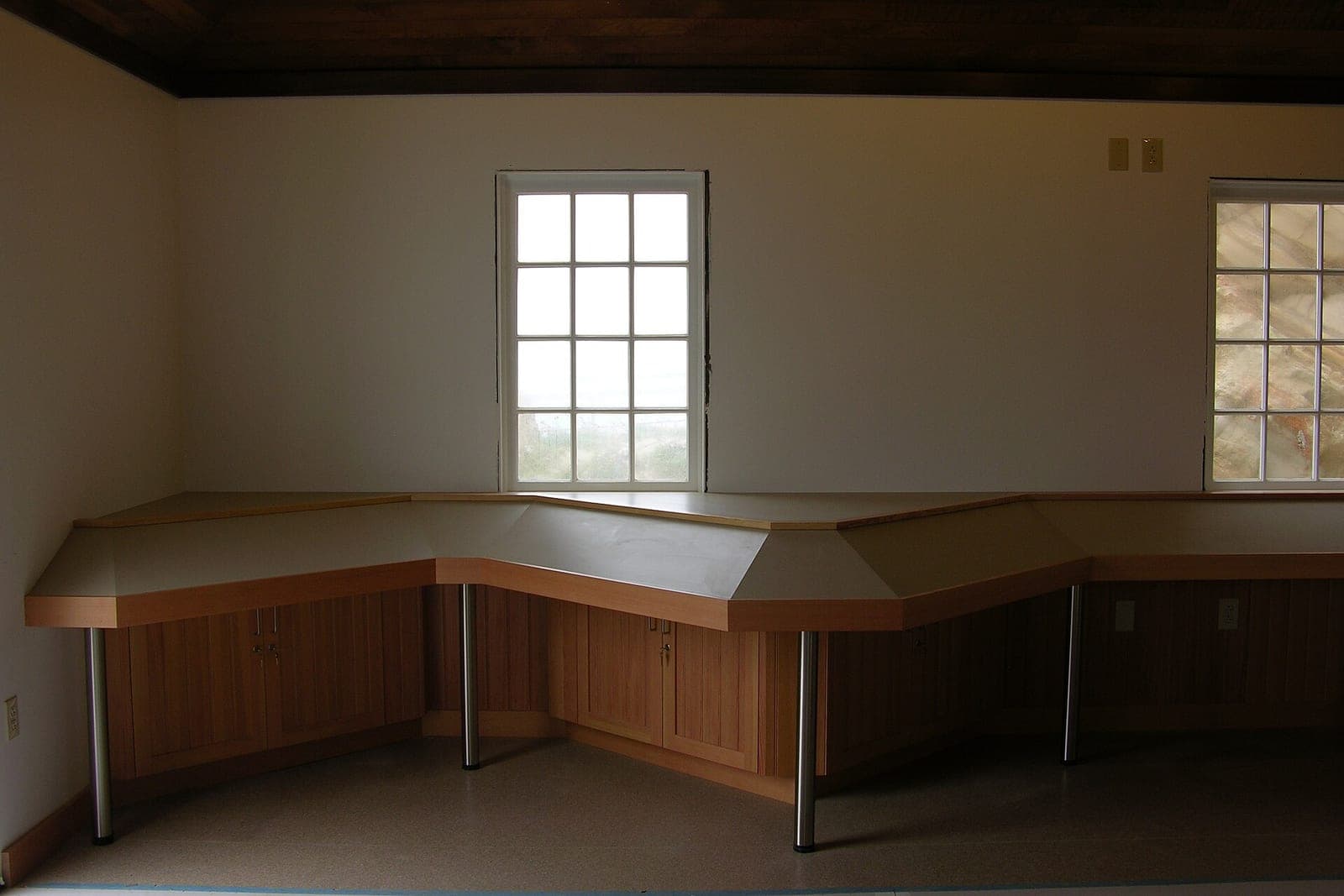 New wooden cabinets and an angled countertop run along the wall of an empty room under two windows