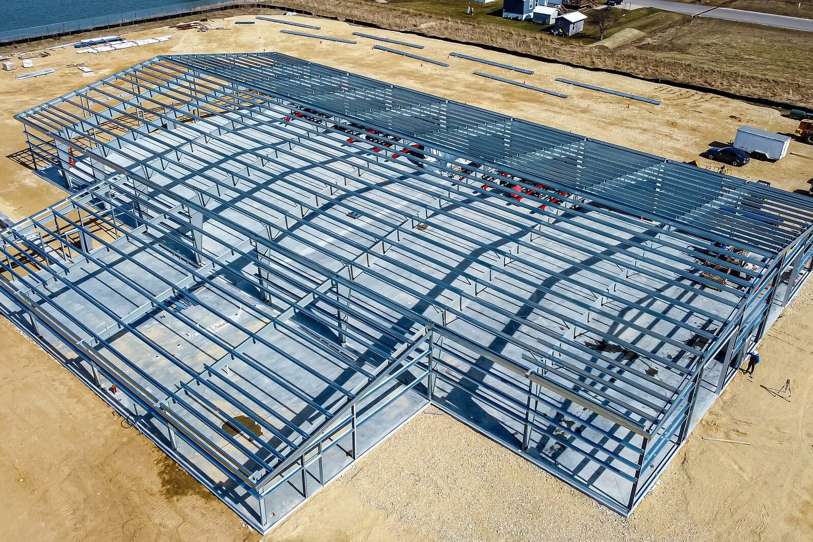 An aerial view of a bare steel building frame rising on a sandy lot beside a grassy field