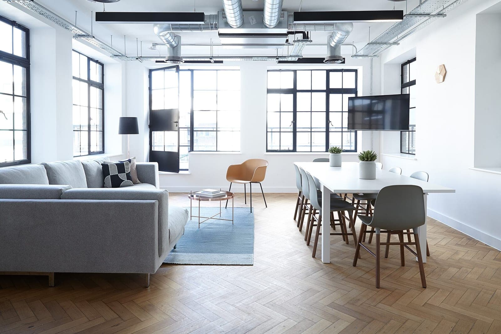A bright office with a gray sofa, a white table with gray chairs and tall black-framed windows over a wood floor