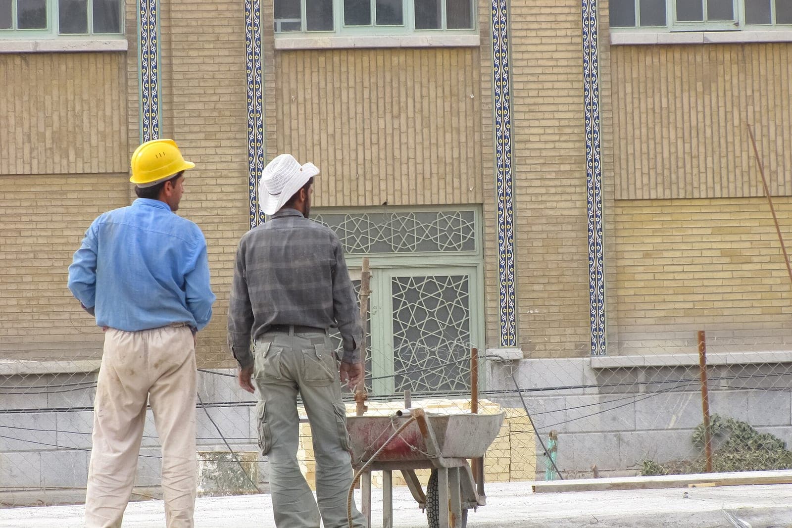 Two workers, one in a yellow hard hat, stand by a wheelbarrow at the wire fence of a job site in front of a tan brick building