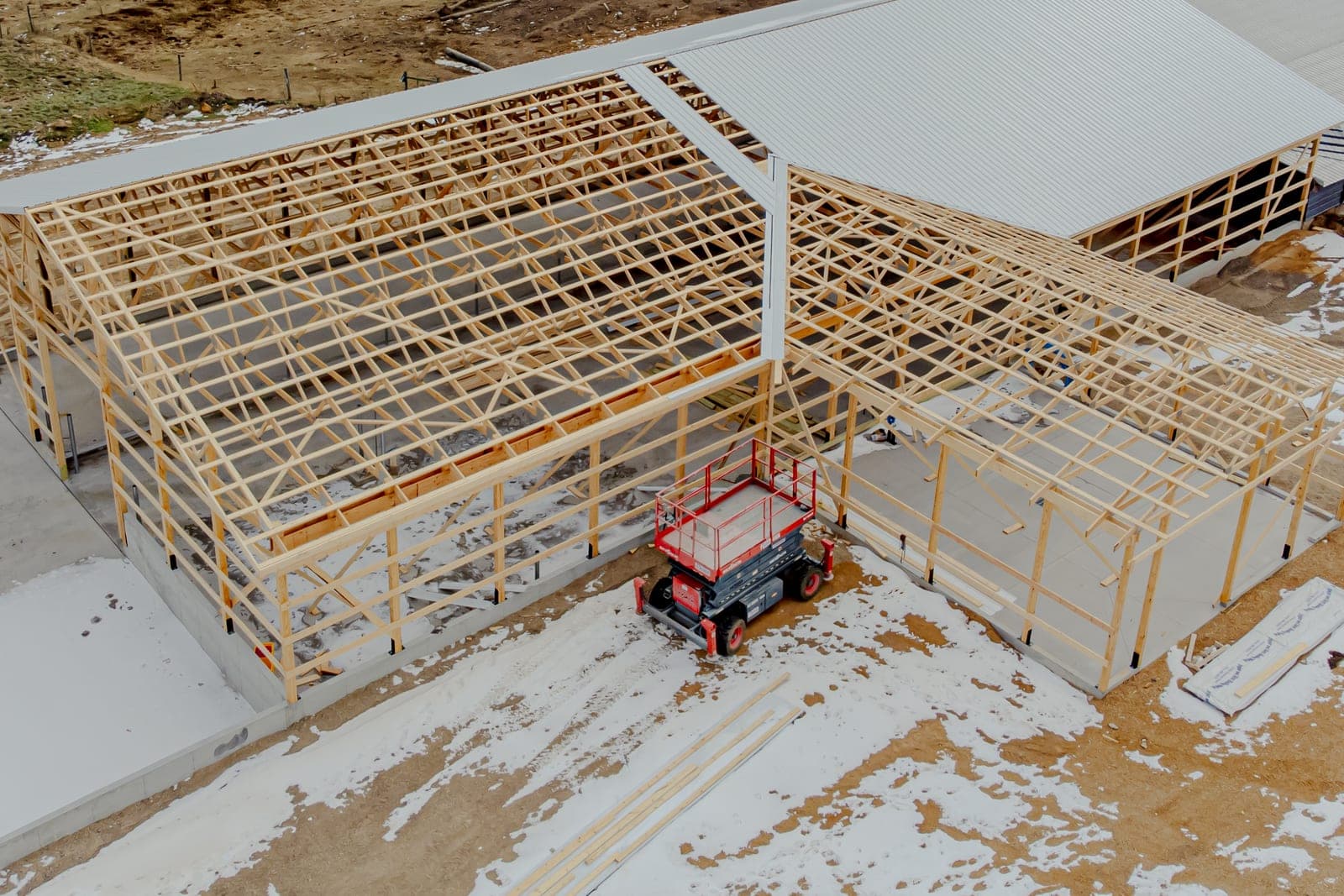 A red scissor lift parked beside the wood frame of a large building under construction, with patches of snow on the ground