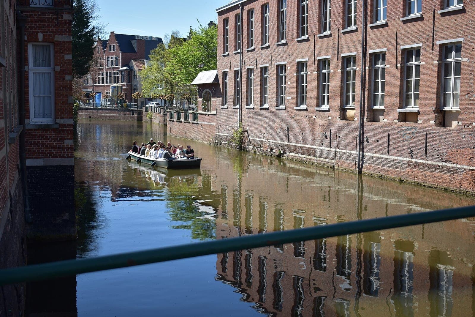 A small open boat full of adult passengers glides along a quiet canal between tall brick buildings