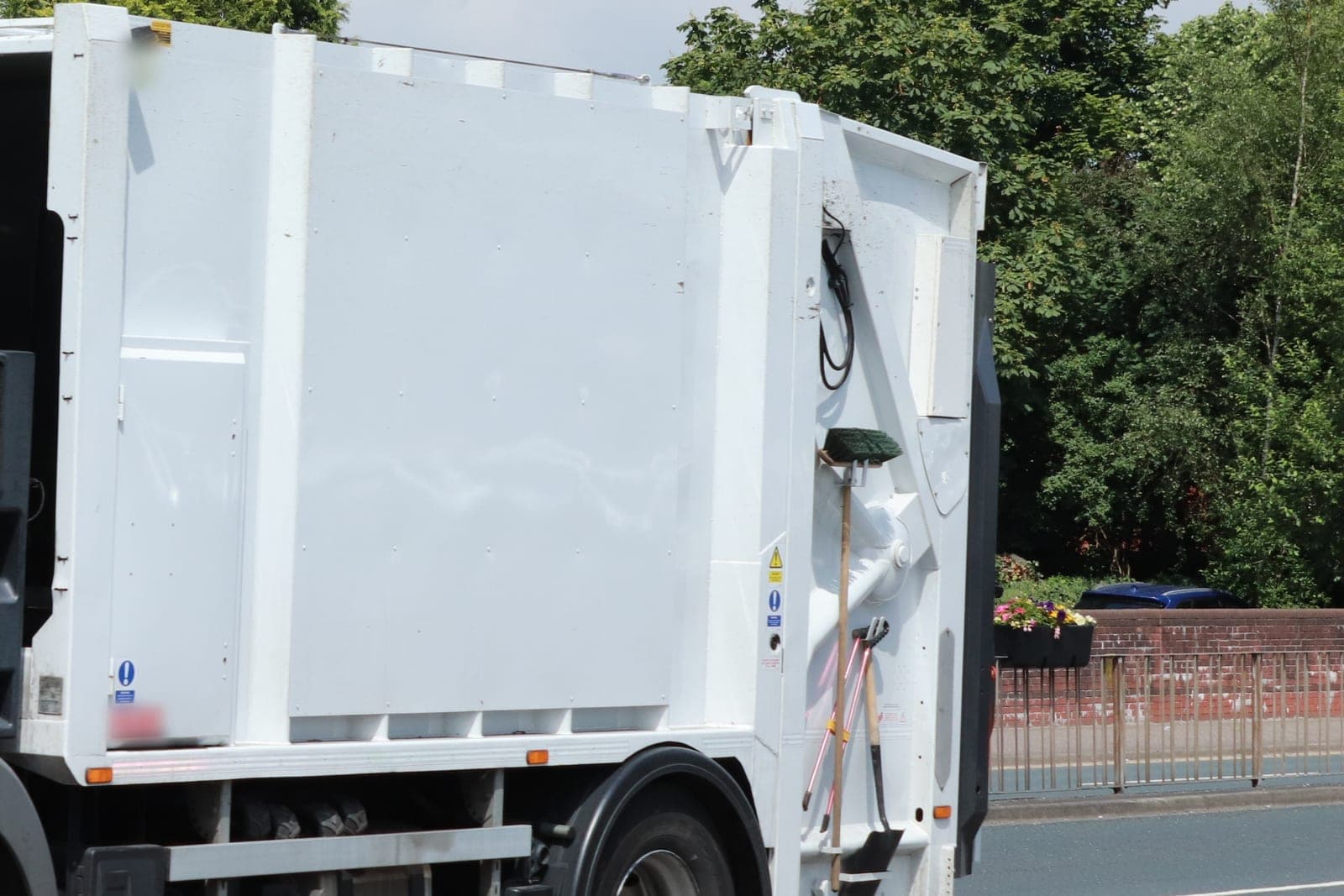 The white compactor body of a trash truck with a broom and shovel clipped to its back