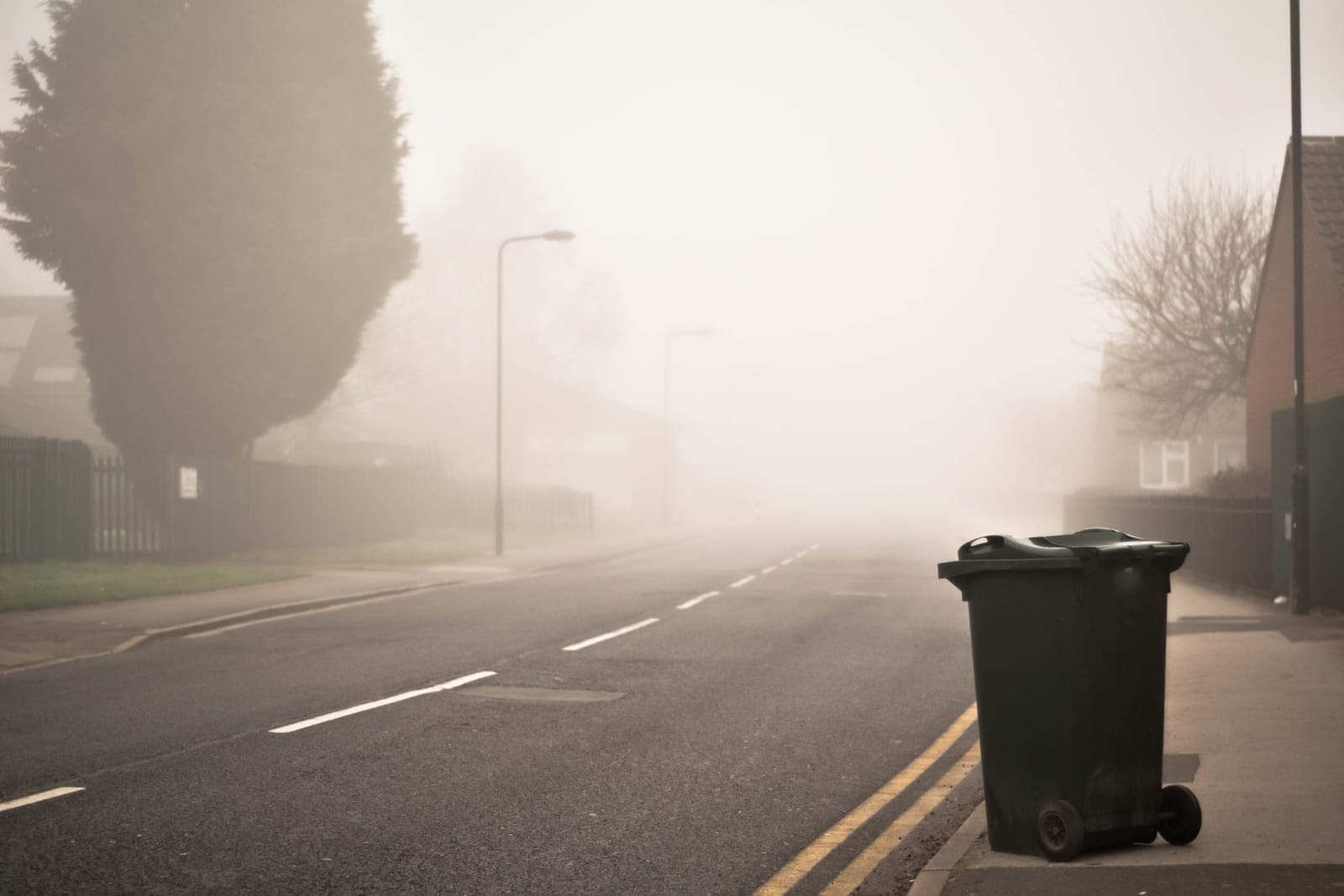 A single dark wheeled trash cart waits at the curb of a foggy, empty street