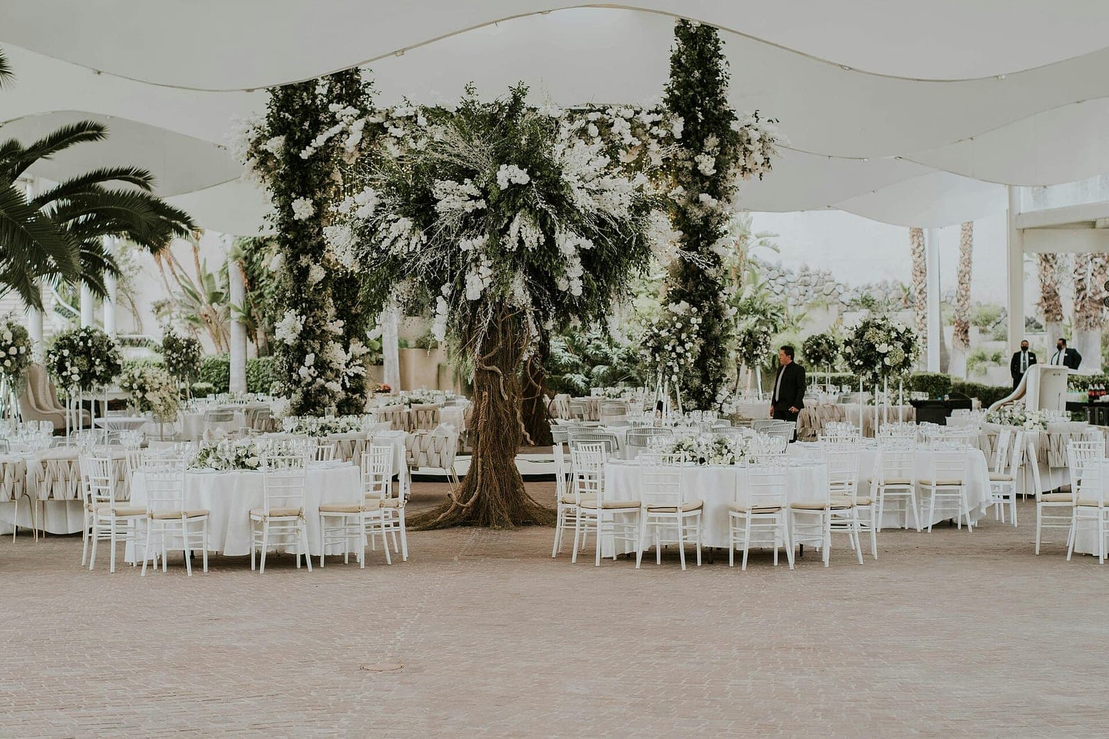 Round tables with white cloths and white chairs under a white fabric canopy, with a tall floral tree in the middle and staff in the distance