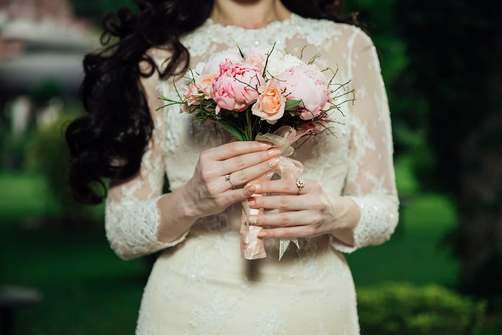 A bride in a lace dress holding a small bouquet of pink peonies and peach roses, seen from the neck down