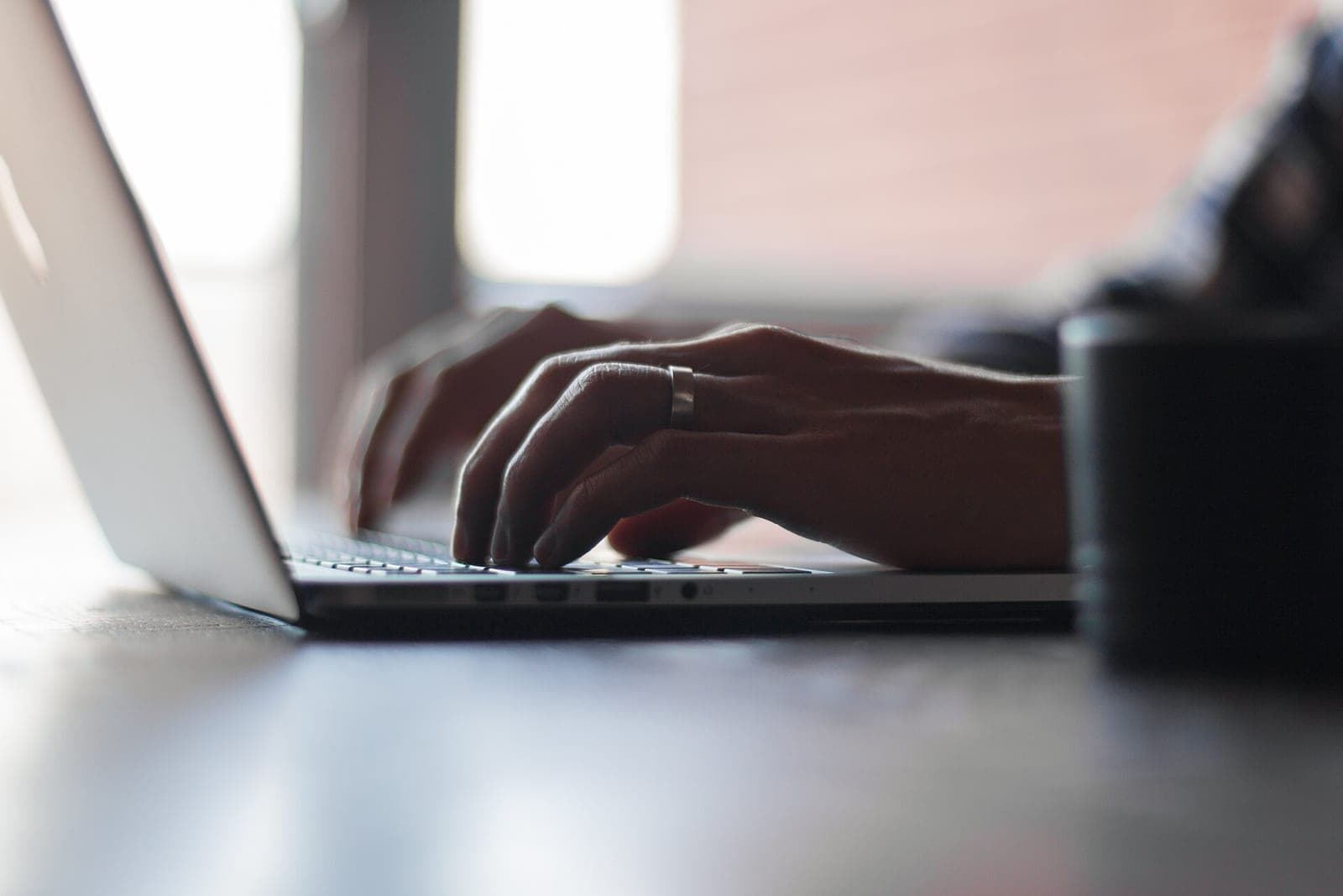 Two hands typing on a silver laptop on a dark desk, with a mug blurred in the foreground