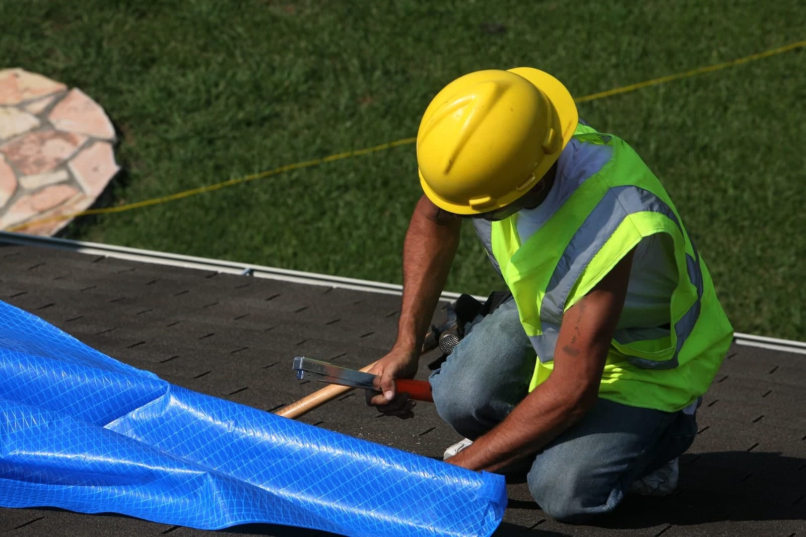 A worker in a yellow hard hat and safety vest fastens a blue tarp on a house roof