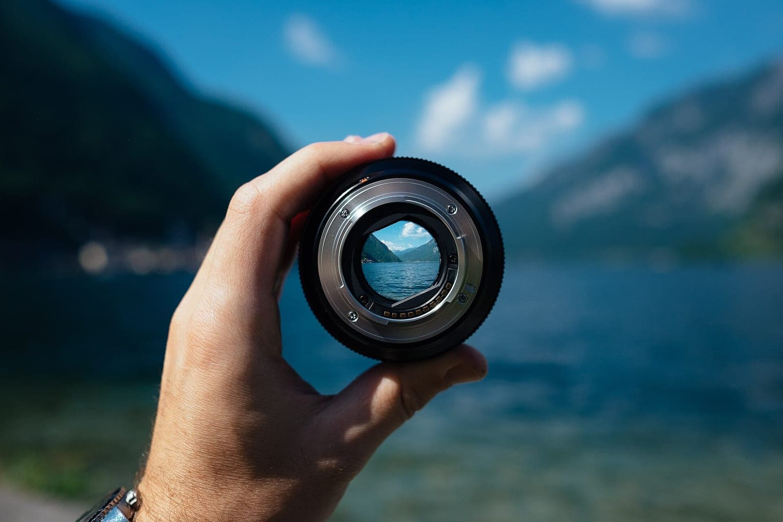 A hand holds up a camera lens that frames a mountain lake under a blue sky