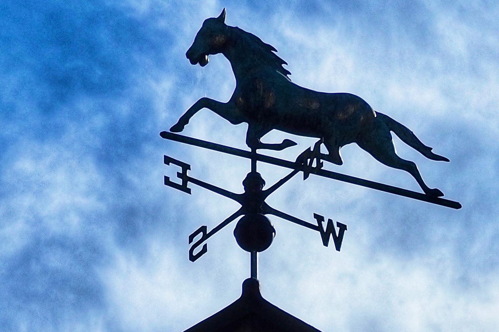A black horse weather vane with compass arms on a rooftop against a cloudy blue sky