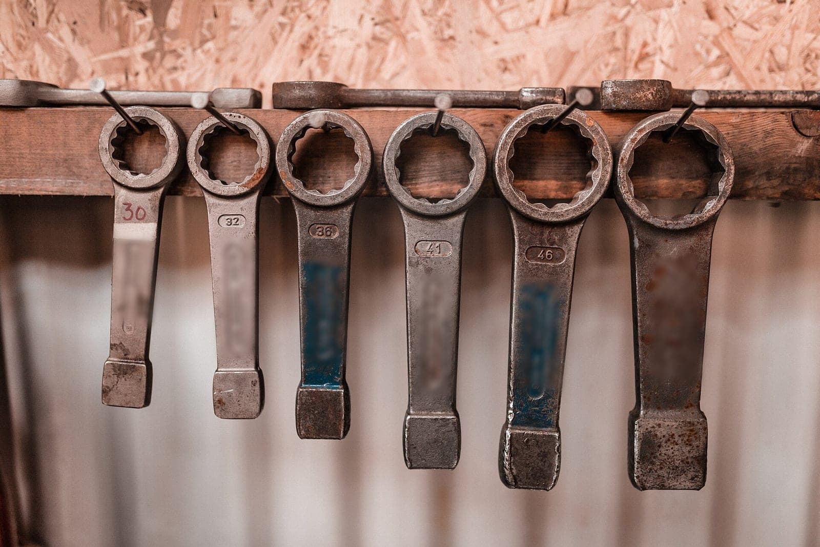 Six worn ring wrenches hanging in size order from nails on a wooden rail