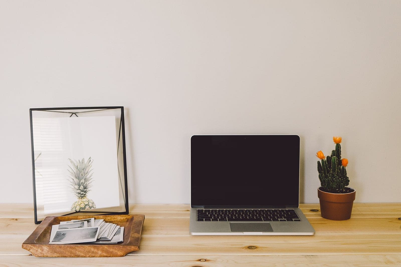 An open laptop with a dark screen on a light wooden table, next to a potted cactus and a framed print of a pineapple